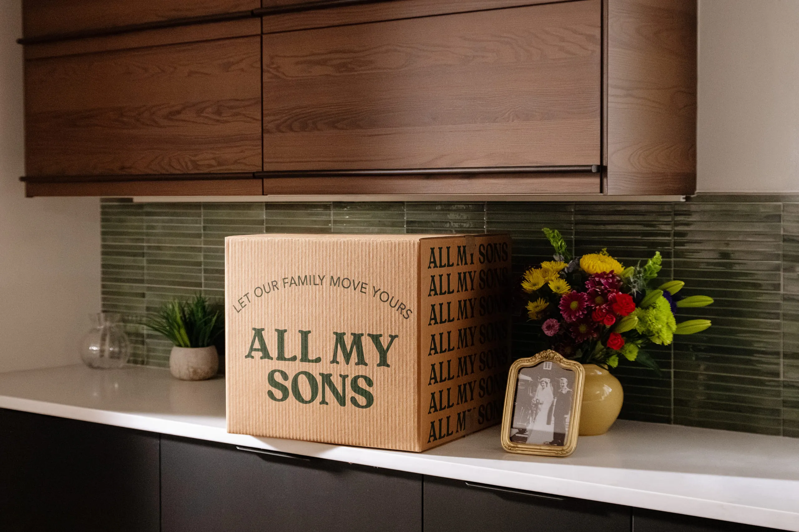 An All My Sons moving box on a kitchen counter beside a vase of flowers and a framed wedding photograph.