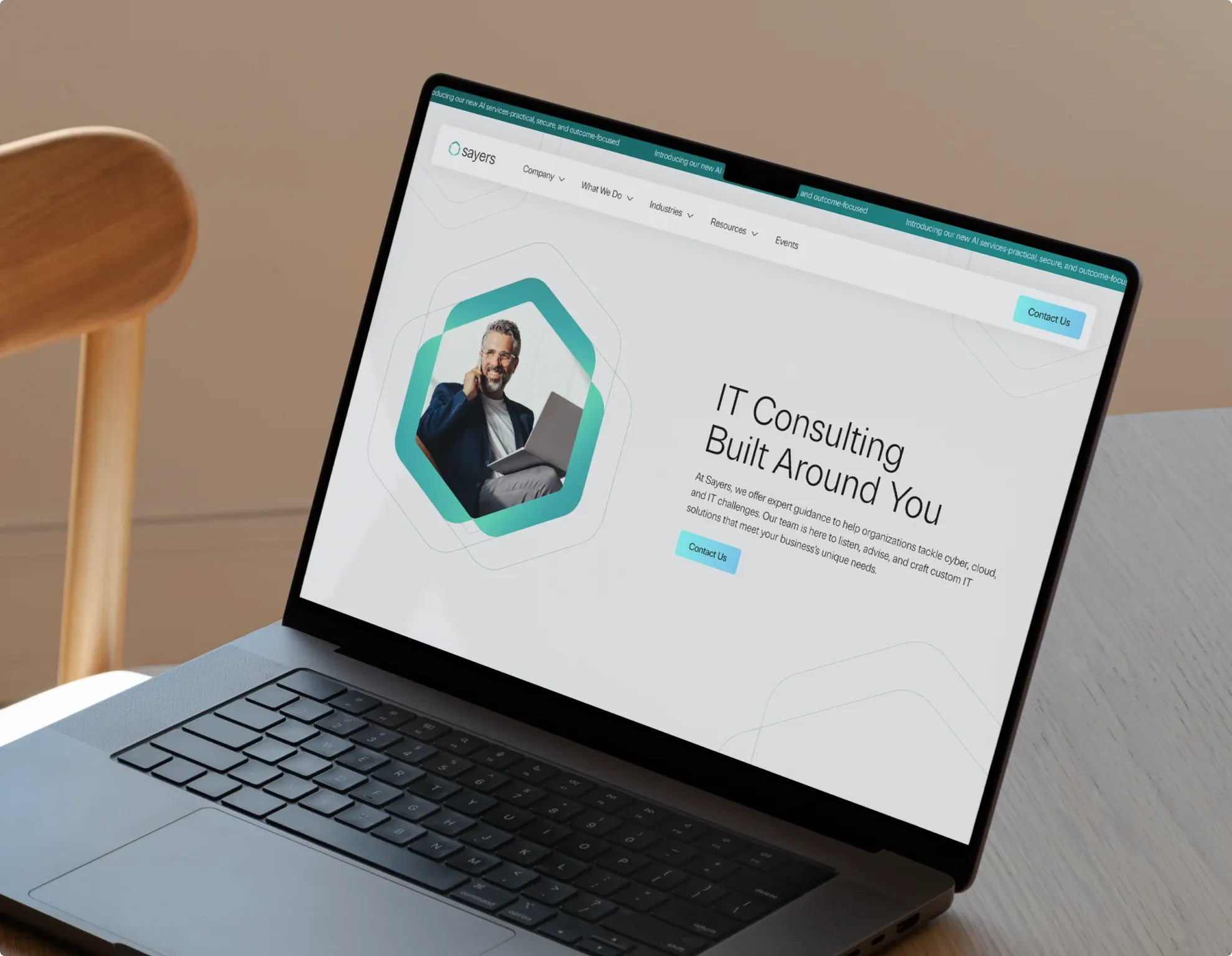 The new Sayers website open on a laptop on a wooden chair, a consultant framed in the hexagon on its homepage.