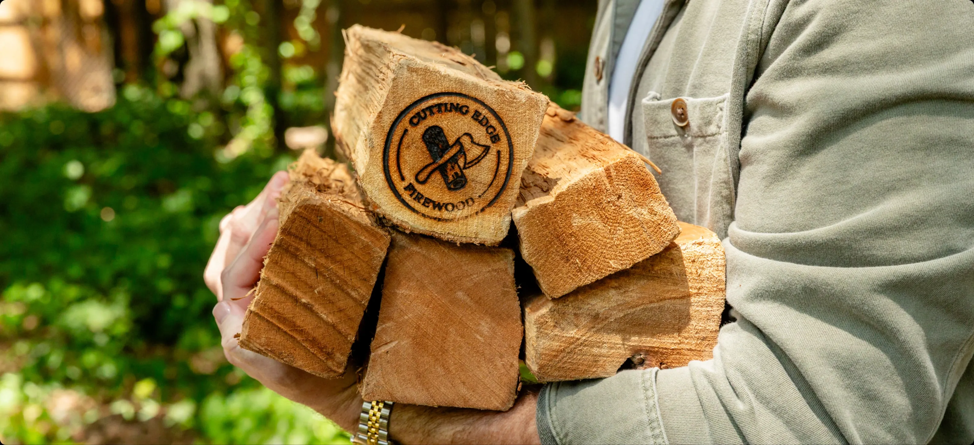 Hands holding an armful of Cutting Edge Firewood logs, the brand burned into one.