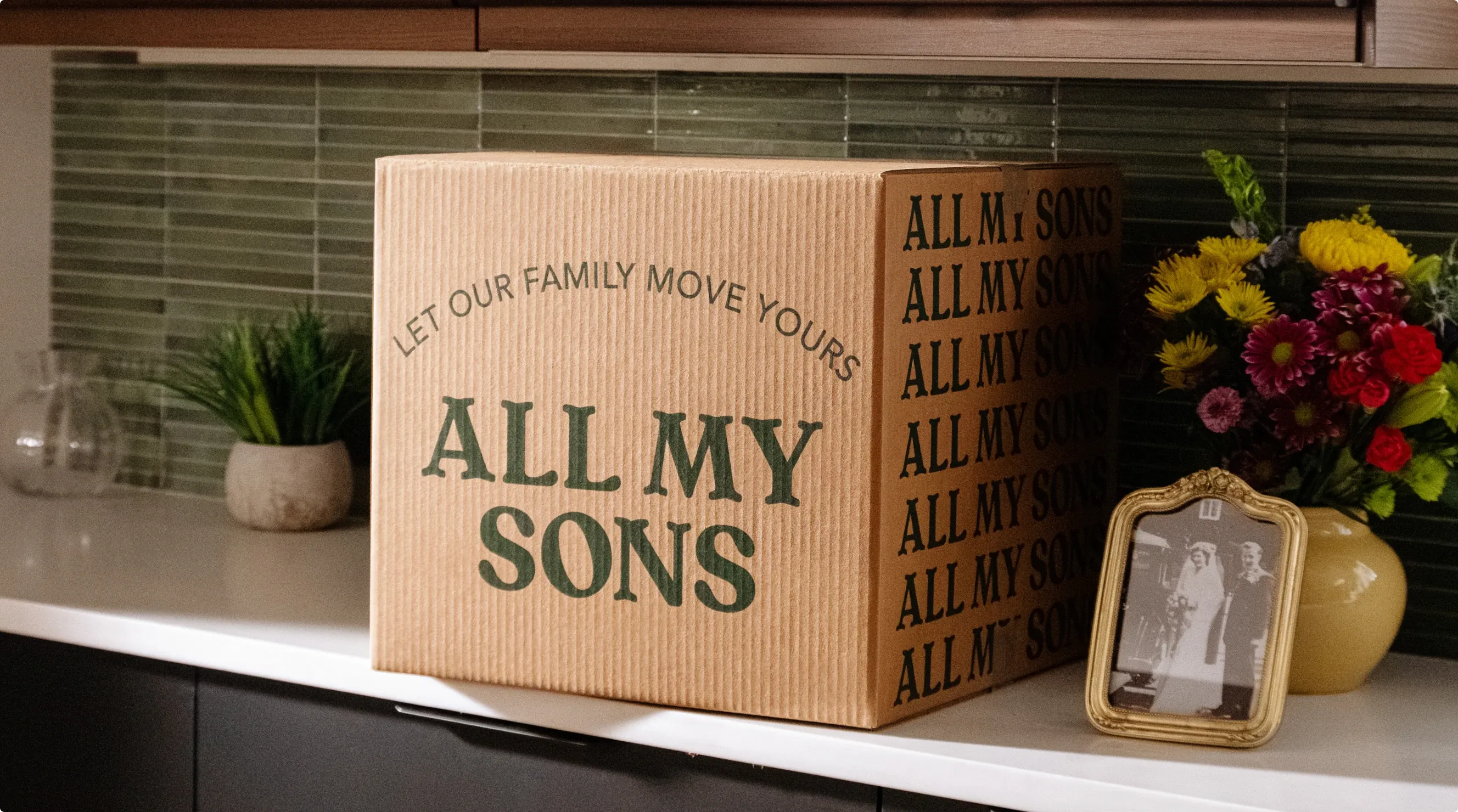 An All My Sons moving box on a kitchen counter beside a vase of flowers and a framed wedding photograph.
