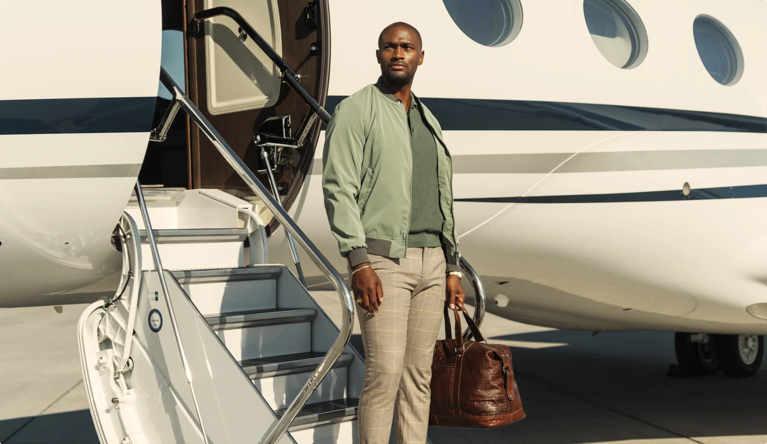 A traveller in a sage jacket stepping down the airstair of a private jet, a leather holdall in hand.