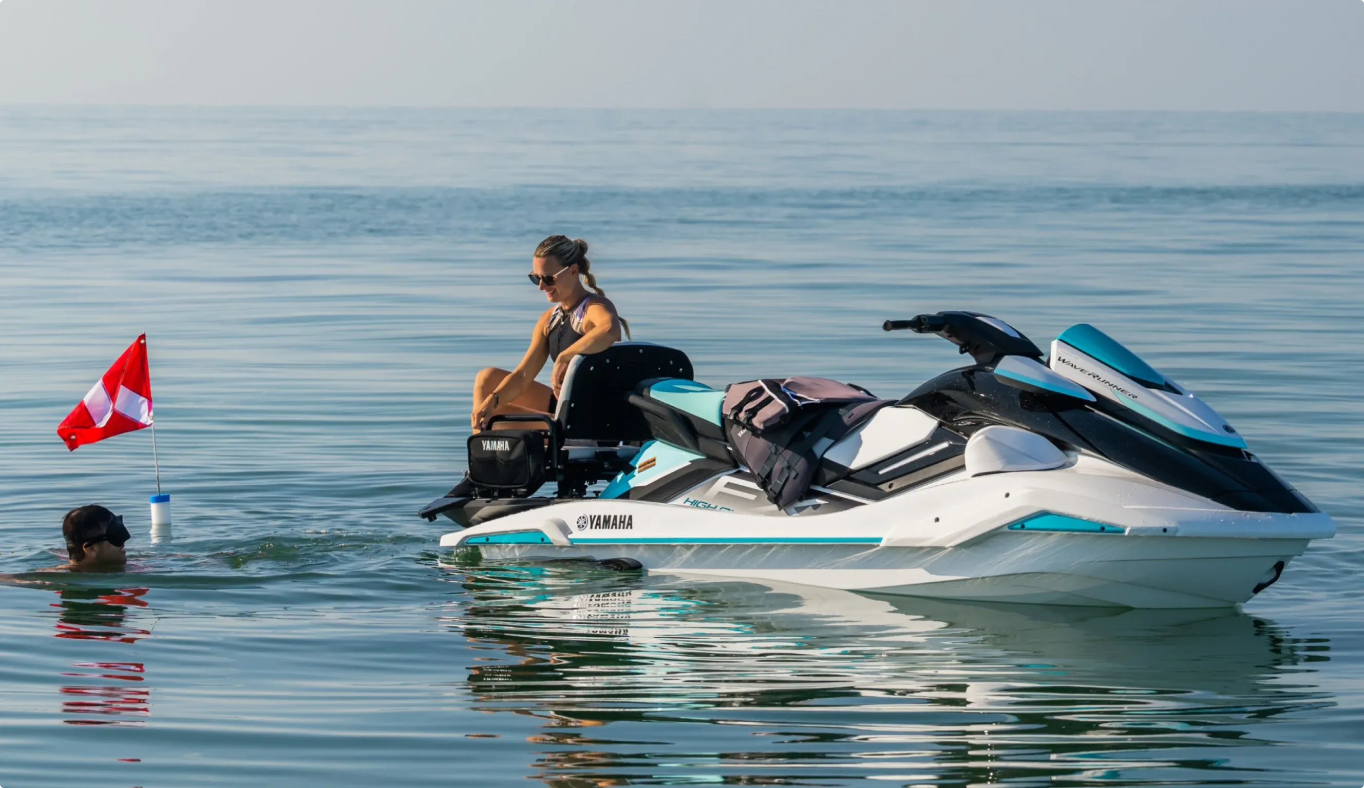 A rider on a Yamaha WaveRunner idling on calm water beside a swimmer and a red marker flag.